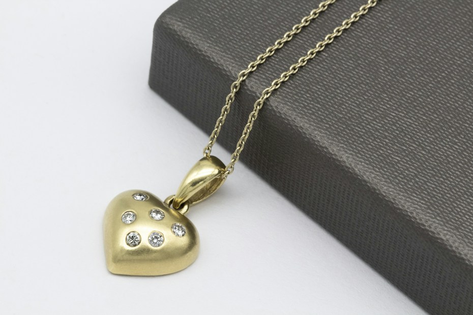A gold heart-shaped pendant adorned with seven small, sparkling diamonds is attached to a delicate chain. The pendant and chain are placed atop a textured, dark gray box on a white surface.