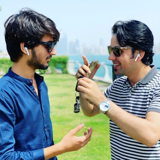 Two men with dark hair wearing sunglasses and AirPods stand in a park. One man is wearing a blue shirt and the other a striped polo shirt. They appear to be engaged in conversation as they look at a phone held by the man in the striped shirt. The background shows a grassy area with hedges and a body of water, suggesting a waterfront location.