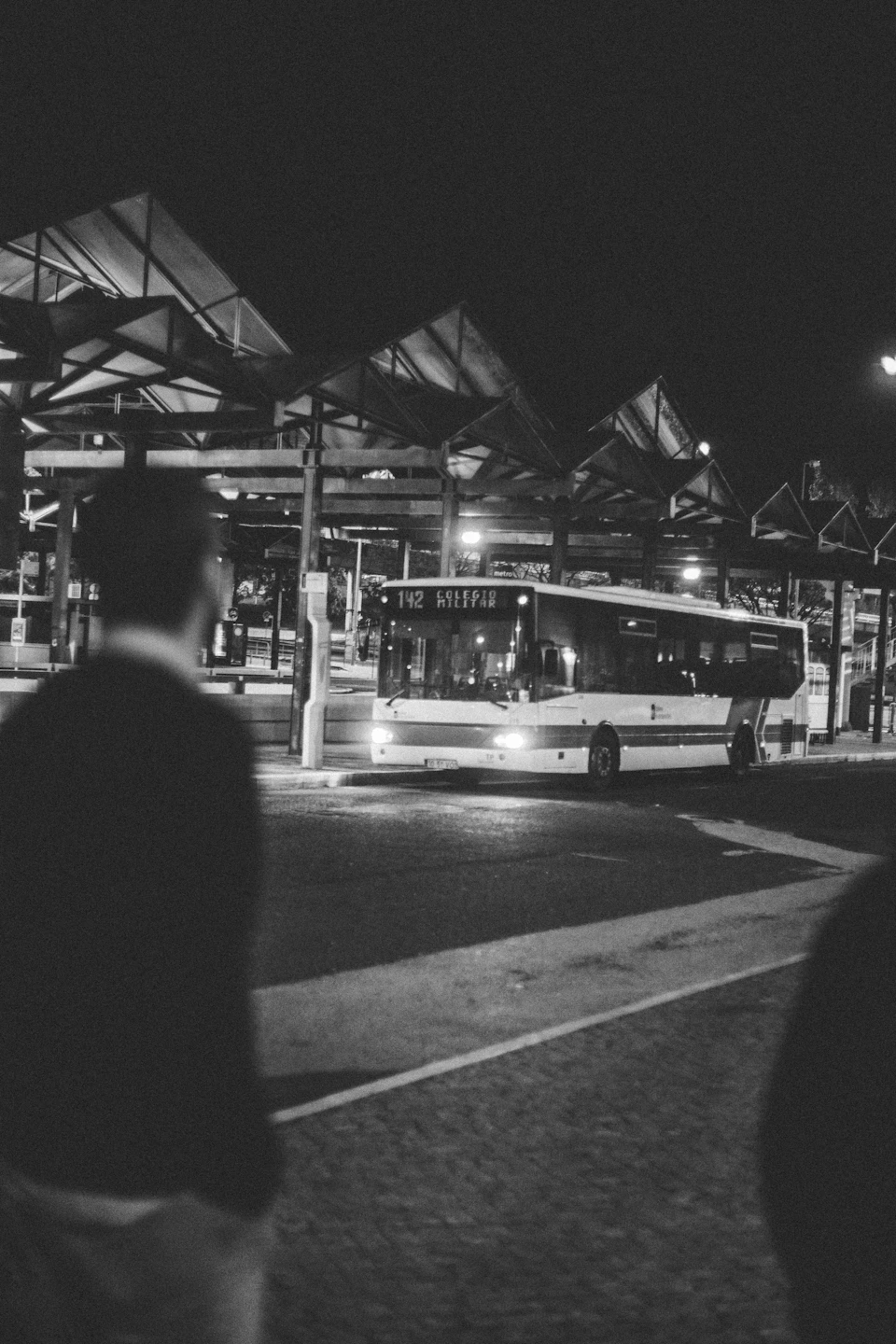 The image depicts a bus stop at night, with a white bus parked under a covered structure and two individuals standing on the sidewalk nearby. The scene is captured from behind one of these individuals, creating an interesting perspective that adds depth to the composition.