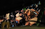 A group of people gathered for an event, likely religious or spiritual in nature. A young boy with a book placed on his head is prominently visible. People around him are holding similar books and some are wearing masks. The setting appears dimly lit with a focus on reading or prayer.