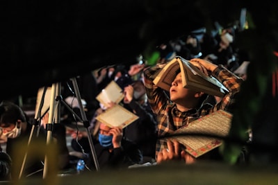 A group of people gathered for an event, likely religious or spiritual in nature. A young boy with a book placed on his head is prominently visible. People around him are holding similar books and some are wearing masks. The setting appears dimly lit with a focus on reading or prayer.