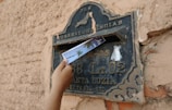A hand is inserting a postcard into an old, rustic metal mailbox mounted on a textured wall. The mailbox is labeled with various inscriptions and numbers, suggesting its location and purpose.