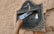A hand is inserting a postcard into an old, rustic metal mailbox mounted on a textured wall. The mailbox is labeled with various inscriptions and numbers, suggesting its location and purpose.