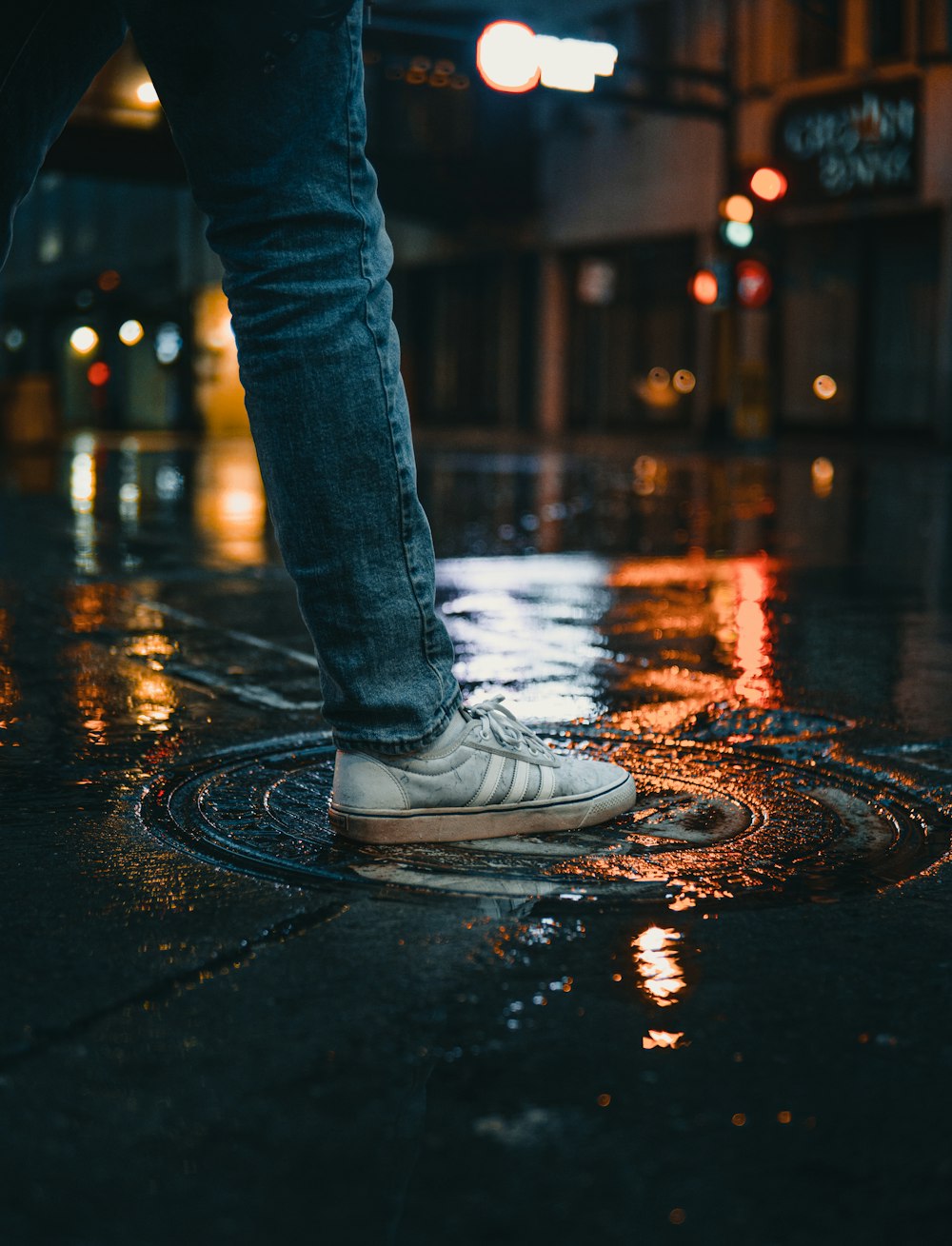 person in blue denim jeans and white sneakers standing on wet road during night time