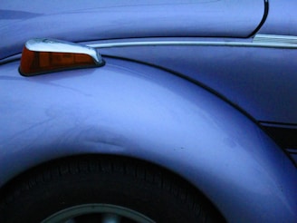 A close-up view of a vintage car's fender is displayed, highlighting its sleek, curved design and metallic violet-blue paint. The image features a chrome side marker with orange glass, which adds a touch of classic automotive style.