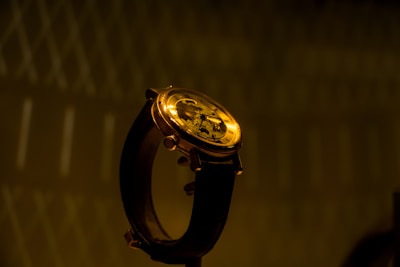 A close-up of an elegant wristwatch with a brown leather strap and a gold-colored, intricate mechanical dial. The background is dark with a subtle geometric pattern, creating a sophisticated and luxurious atmosphere.