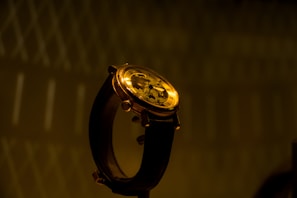 A close-up of an elegant wristwatch with a brown leather strap and a gold-colored, intricate mechanical dial. The background is dark with a subtle geometric pattern, creating a sophisticated and luxurious atmosphere.