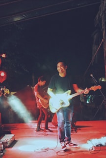 A band is performing on stage, featuring a guitarist who is smiling and seems to be enjoying the music. The scene is lively with bright stage lights and a smokey atmosphere, creating an energetic concert vibe.