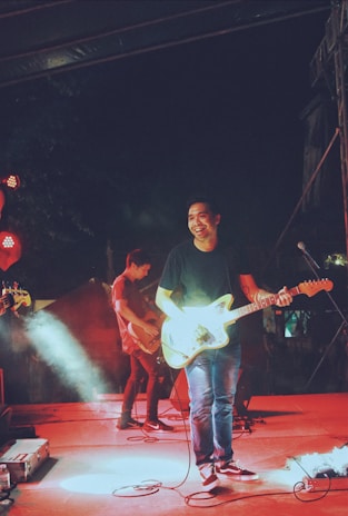 A band is performing on stage, featuring a guitarist who is smiling and seems to be enjoying the music. The scene is lively with bright stage lights and a smokey atmosphere, creating an energetic concert vibe.