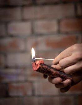 A lit match is being used to ignite a red firecracker held by a hand. The background features a brick wall with a blurred effect.