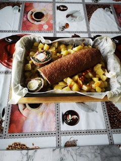 A rectangular tray filled with cooked diced potatoes and a rolled meat dish sliced to reveal stuffing, resting on a decorative tablecloth featuring coffee and food-themed images.