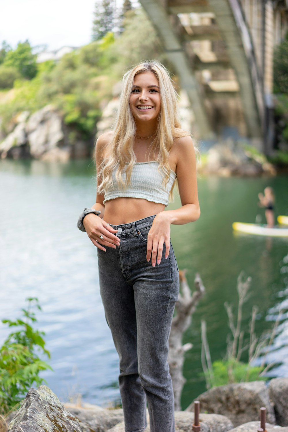 woman in white tank top and blue denim jeans standing near body of water during daytime