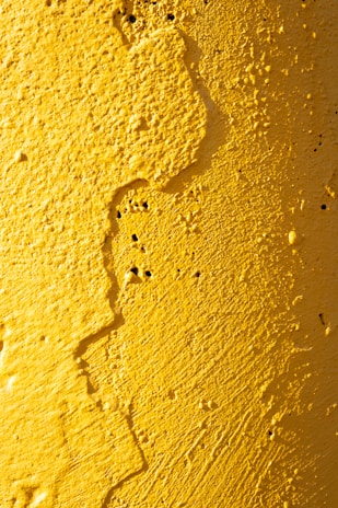 A textured surface covered with yellow paint, featuring rough patches and small indents. The paint appears to have been applied thickly, creating an uneven and rugged texture.