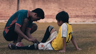 A man kneels on the grass, helping a young boy with a soccer uniform who is sitting on the ground. The interaction suggests care and attentiveness, with both individuals focused on adjusting the boy's shin guard or soccer sock. A blurred brick wall is visible in the background.
