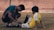 A man kneels on the grass, helping a young boy with a soccer uniform who is sitting on the ground. The interaction suggests care and attentiveness, with both individuals focused on adjusting the boy's shin guard or soccer sock. A blurred brick wall is visible in the background.