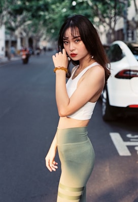 A woman with long dark hair stands confidently on a city street. She is wearing a white tank top and olive green leggings, accessorized with a beaded bracelet on her wrist. In the background, a white car is parked, and trees line the street, casting soft shadows.