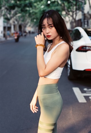 A woman with long dark hair stands confidently on a city street. She is wearing a white tank top and olive green leggings, accessorized with a beaded bracelet on her wrist. In the background, a white car is parked, and trees line the street, casting soft shadows.