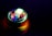 A spinning top emits vibrant colors of light as it rotates on a dark surface. The light creates a circular, almost hypnotic pattern with shades of green, blue, red, and purple reflecting on the surface.