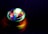 A spinning top emits vibrant colors of light as it rotates on a dark surface. The light creates a circular, almost hypnotic pattern with shades of green, blue, red, and purple reflecting on the surface.