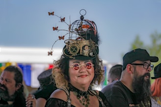A person wearing a steampunk-inspired outfit with a unique hat adorned with gears, a small birdcage, and decorative butterflies. The individual is smiling and wearing red-tinted round glasses in a crowd. The background features blurry figures and colorful flags in front of a clear blue sky.