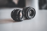 Two camera lenses are placed on a smooth surface. The lenses have metallic and glass components, and visible apertures. The lighting is soft, creating a gentle reflection on the surface.
