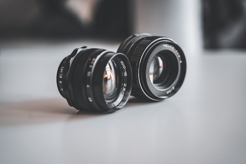 Two camera lenses are placed on a smooth surface. The lenses have metallic and glass components, and visible apertures. The lighting is soft, creating a gentle reflection on the surface.