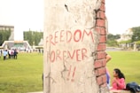 A concrete pillar with the words 'FREEDOM FOREVER!!' written in red graffiti. The pillar is weathered, with visible cracks and exposed reddish bricks. In the background, a group of people is gathered on a green lawn, with trees and a building visible.