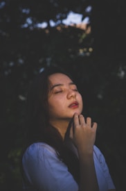 A woman is depicted with her eyes closed, basking in sunlight that highlights her face. Her long dark hair flows naturally, and she wears a light-colored shirt. Her hand is gently touching her chin, and she appears serene and peaceful. The background is dark, likely consisting of blurred foliage.