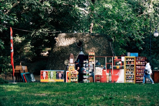 A small kiosk set up outdoors with various snacks and drinks displayed on shelves. The kiosk is positioned in front of a large tree, providing a natural backdrop. Various colorful packages, cans, and bottles are visible, indicating a variety of items for sale. The area is surrounded by lush green foliage, suggesting a park or garden setting. A person walks by the kiosk, dressed casually.