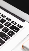 A close-up view of a laptop keyboard, focusing on keys such as delete, return, and shift. The keyboard features black keys with white lettering and is surrounded by a white frame. In the bottom right corner, a partial view of a white object with an orange accent is visible.