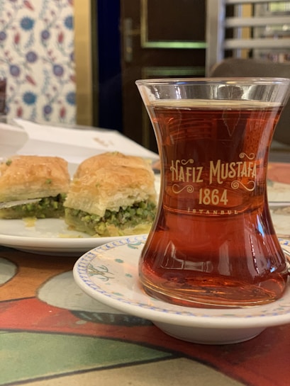 A glass of Turkish tea with the text 'Hafiz Mustafa 1864' on it, placed on a decorated saucer. In the background, there is a white plate with pieces of pistachio baklava. The setting appears to be a traditional caf&eacute;, indicated by the ornate wallpaper and a wooden door.
