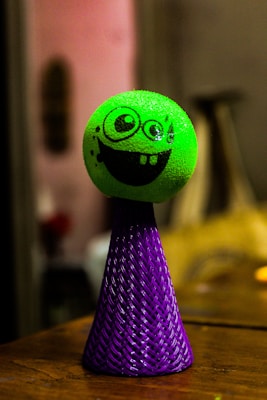 A toy featuring a bright green, textured sphere with a cartoonish, black smiling face is positioned on top of a woven, cone-shaped base in vibrant purple. The setting appears to be indoors with soft, warm lighting, showing some out-of-focus background elements.