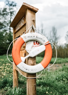 A life preserver ring with text 'Uppsala Kommun Trygg-Hansa' is mounted on a wooden signpost outdoors. Surrounding the signpost is a grassy area with some trees and a cloudy sky in the background.