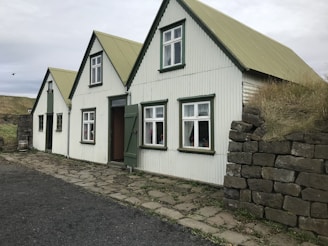 A cozy house with a corrugated metal exterior and gabled roofs, featuring mossy rock walls and traditional windows. The house blends harmoniously into a rustic landscape with overcast skies.