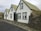 A cozy house with a corrugated metal exterior and gabled roofs, featuring mossy rock walls and traditional windows. The house blends harmoniously into a rustic landscape with overcast skies.