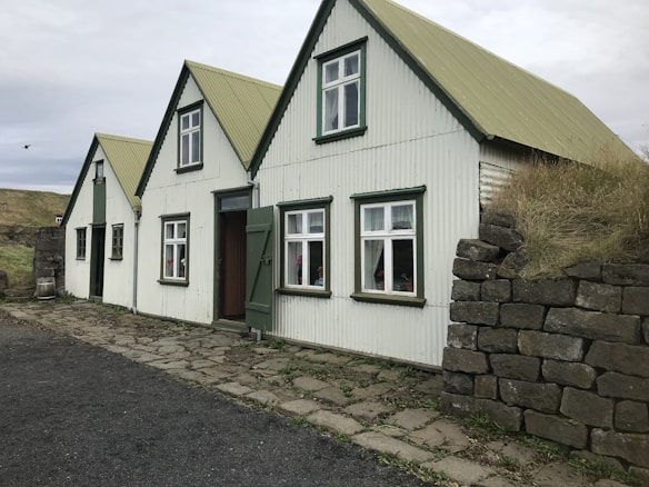 A cozy house with a corrugated metal exterior and gabled roofs, featuring mossy rock walls and traditional windows. The house blends harmoniously into a rustic landscape with overcast skies.