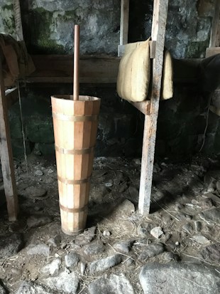 A rustic interior with a wooden churn placed on a stone floor. The walls are made of dark, mossy stones, and there are wooden beams supporting burlap sacks hung from a rack. The setting has an old, traditional look, resembling a barn or cellar environment.