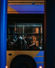 A city bus is seen through the window with passengers sitting inside. The image is taken during nighttime, creating a dimly lit atmosphere. The bus has a distinctive yellow and blue color scheme. Passengers are wearing face masks and are seated on the bus, some looking at their phones.