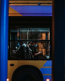 A city bus is seen through the window with passengers sitting inside. The image is taken during nighttime, creating a dimly lit atmosphere. The bus has a distinctive yellow and blue color scheme. Passengers are wearing face masks and are seated on the bus, some looking at their phones.