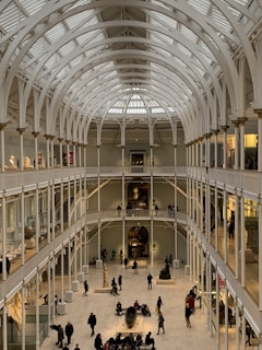 A large, open interior space of a museum with a high, arched ceiling made of white metal beams. There are multiple levels with balconies overlooking a central area that contains several people walking and observing displays. The lighting is soft, creating a warm ambiance in the spacious hall.