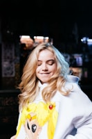 A person with long, blonde hair is wearing a white hoodie featuring an anime character design. The background is dimly lit with some blurred lights, suggesting an indoor setting. The person has a lighthearted expression with closed eyes and a slight smile.