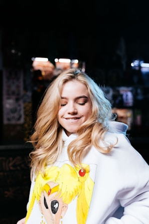 A person with long, blonde hair is wearing a white hoodie featuring an anime character design. The background is dimly lit with some blurred lights, suggesting an indoor setting. The person has a lighthearted expression with closed eyes and a slight smile.