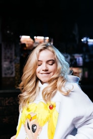 A person with long, blonde hair is wearing a white hoodie featuring an anime character design. The background is dimly lit with some blurred lights, suggesting an indoor setting. The person has a lighthearted expression with closed eyes and a slight smile.