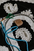An ornate textile piece featuring intricate white embroidery on a dark background. A string of blue beads with green tassels lies on top, along with a small, carved, rectangular stone.