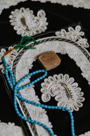 An ornate textile piece featuring intricate white embroidery on a dark background. A string of blue beads with green tassels lies on top, along with a small, carved, rectangular stone.