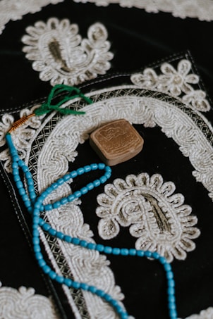 An ornate textile piece featuring intricate white embroidery on a dark background. A string of blue beads with green tassels lies on top, along with a small, carved, rectangular stone.