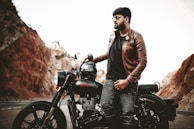A person wearing a brown leather jacket and sunglasses is leaning against a motorcycle parked on a road. The background features rocky terrain with light brown and reddish hues, suggesting a scenic, rugged landscape.