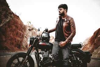 A person wearing a brown leather jacket and sunglasses is leaning against a motorcycle parked on a road. The background features rocky terrain with light brown and reddish hues, suggesting a scenic, rugged landscape.