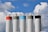 Five tall industrial silos stand against a backdrop of a blue sky with fluffy, white clouds. Each silo is topped with guardrails, and they have different colored bands at the top: red, beige, light blue, gray, and black.