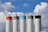 Five tall industrial silos stand against a backdrop of a blue sky with fluffy, white clouds. Each silo is topped with guardrails, and they have different colored bands at the top: red, beige, light blue, gray, and black.
